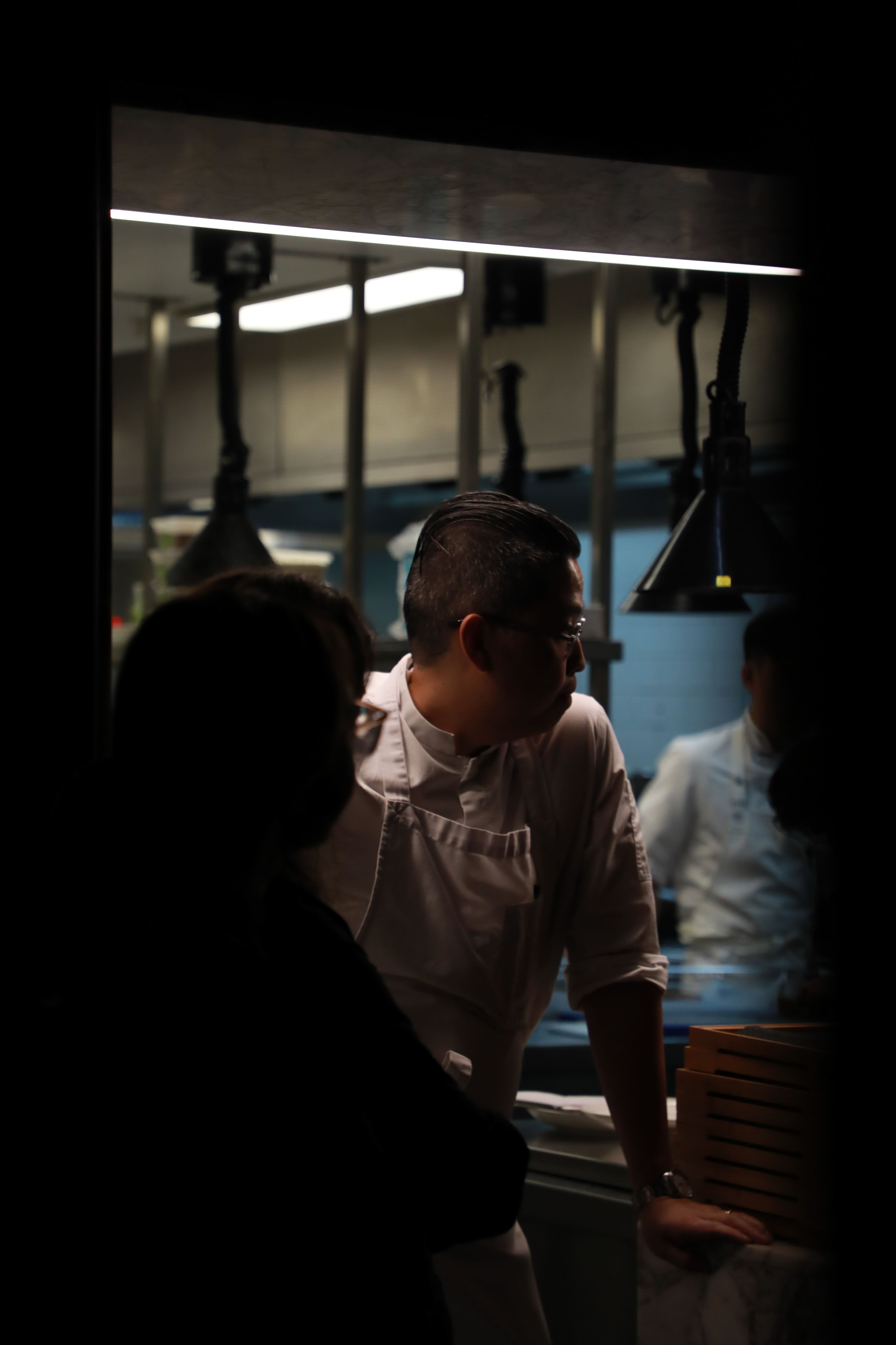 Chef in white uniform leaning over a counter in a dimly lit kitchen with hanging lamps in the background.