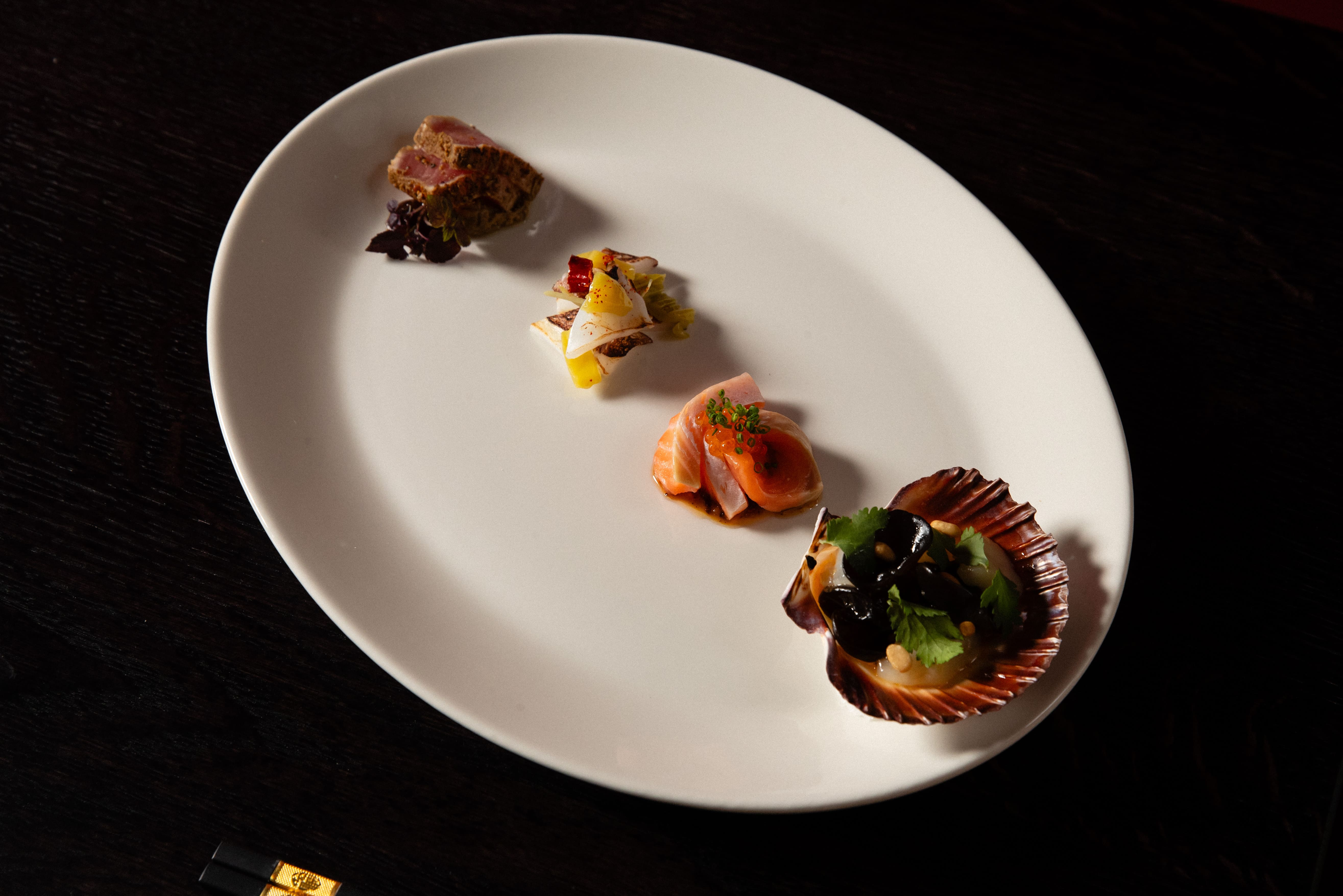 Platter of small tapas on a white plate: a salmon canapé with roe, a layered bite with yellow and white elements, and a seared meat/vegetable piece, arranged on a dark wooden table.