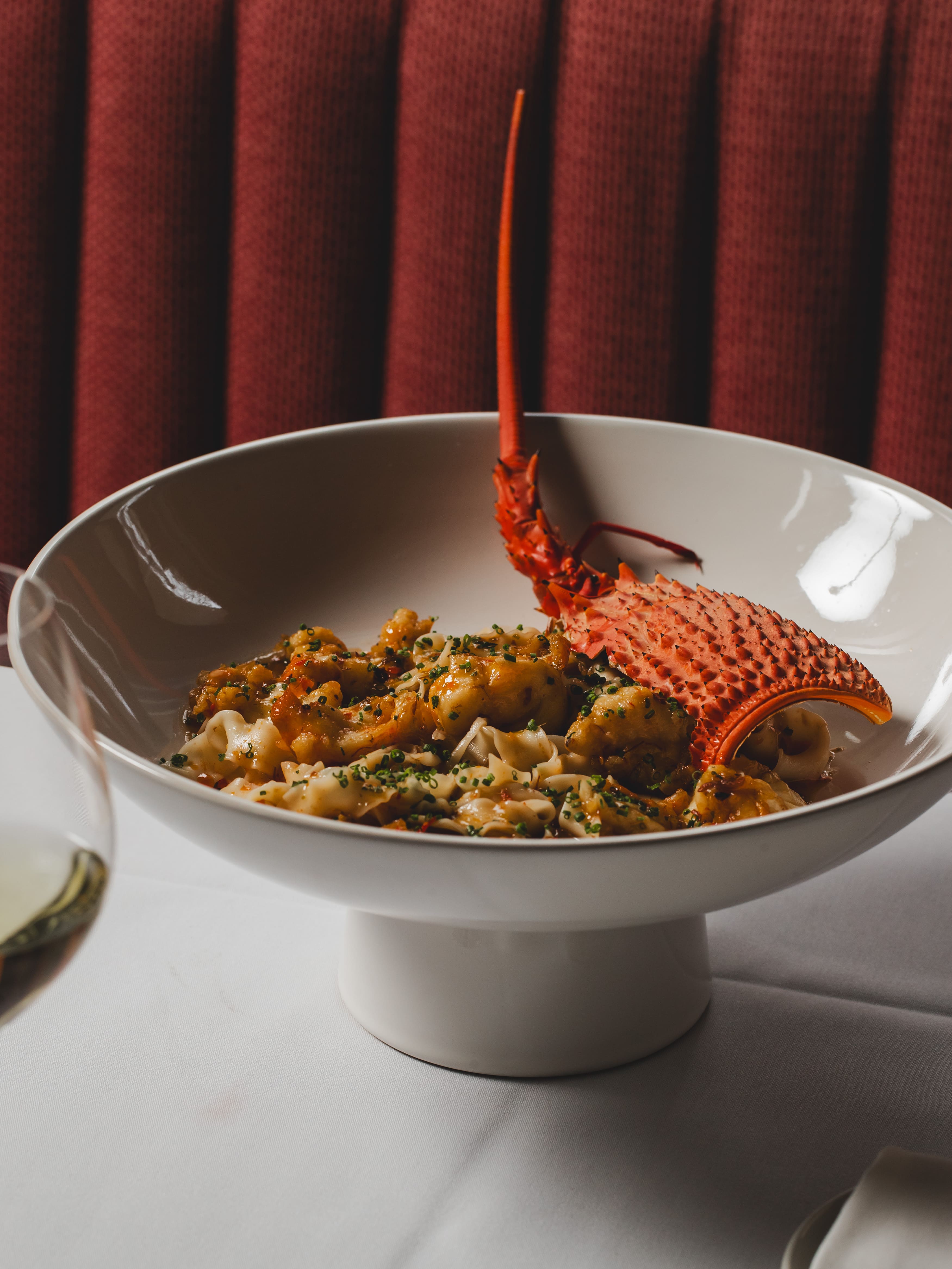 Boiled lobster tail served on pasta in a white bowl, garnished with herbs, against a red curtain backdrop.